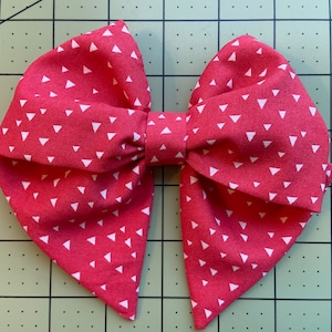 Puede incluir: Un gran lazo rojo con un patrón de triángulos blancos. El lazo está hecho de tela y está atado en forma de lazo clásico.