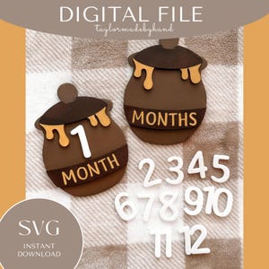May include: A brown honey pot shaped monthly milestone marker with the text "1 month" and a white number "1".  There are also white numbers 2 through 12 for a baby's first year.