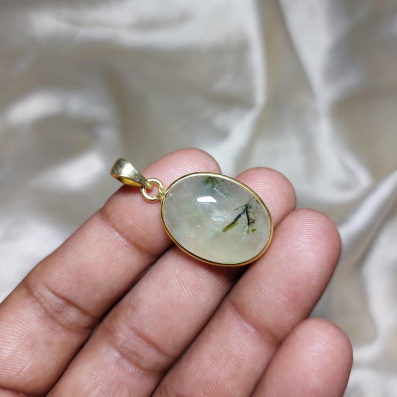 May include: Oval pendant with a gold-coloured frame. The pendant features a light green stone with dark green and black inclusions. The bail is also gold-coloured. The pendant is displayed against a soft, off-white background.