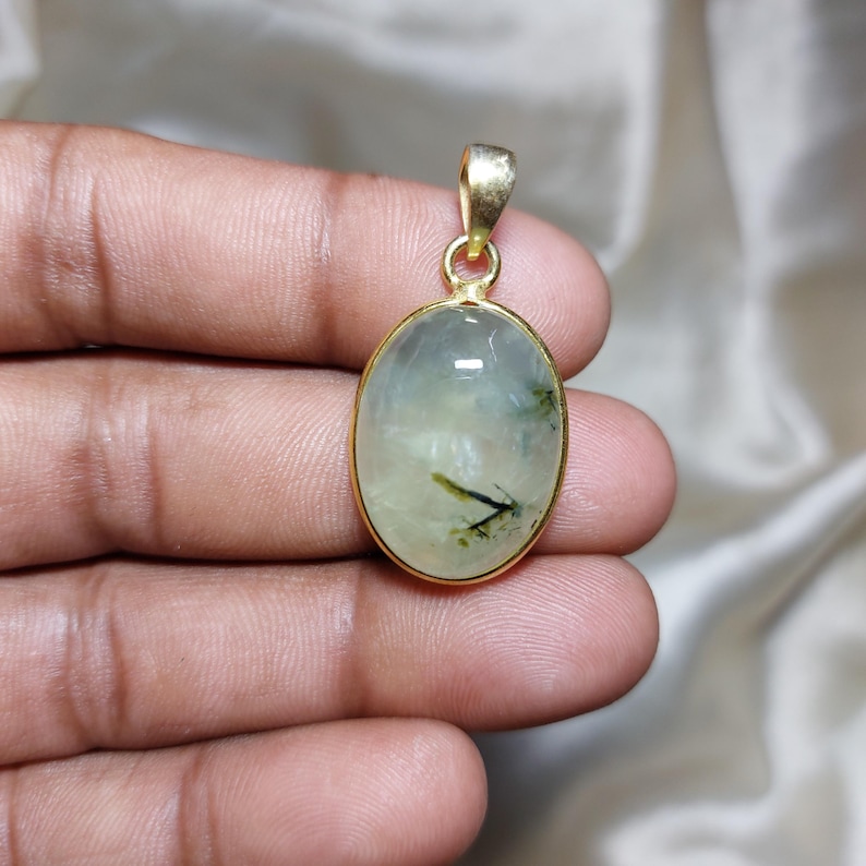 May include: Oval pendant with a gold-coloured frame. The pendant features a light green stone with dark green inclusions. The bail is also gold-coloured. The pendant is held in a person's hand.