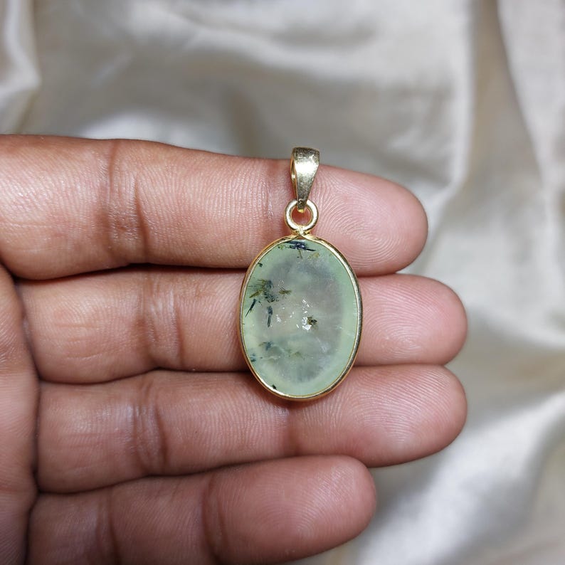 May include: Oval pendant with a pale green gemstone, with dark green inclusions, set in a gold-coloured metal frame. The pendant has a small loop at the top for a chain. The gemstone is approximately 2.5 cm in length.