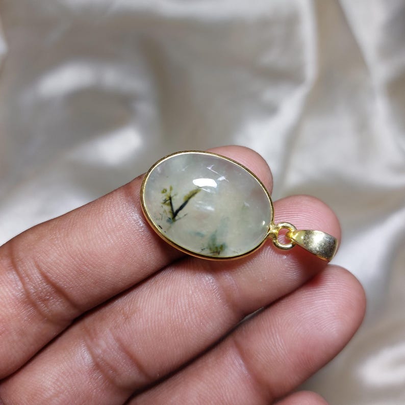 May include: Oval pendant with a gold-coloured frame. The pendant features a translucent, light green stone with dark green and black inclusions. The bail is also gold-coloured. The pendant is held in a hand.