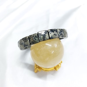 Puede incluir: Una pulsera de pirita con cuentas rectangulares descansa sobre una bola de cristal amarilla. Las cuentas grises metálicas de la pulsera tienen un aspecto texturizado. La bola de cristal se encuentra sobre un pequeño soporte dorado, todo sobre un fondo blanco.