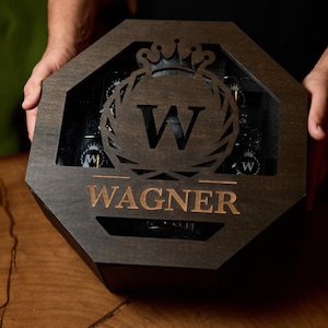 May include: Dark wooden box with an octagonal shape, holding several bottles. The box features a decorative emblem with the letter "W" and the name "Wagner" in a copper color. The box is held by a person.