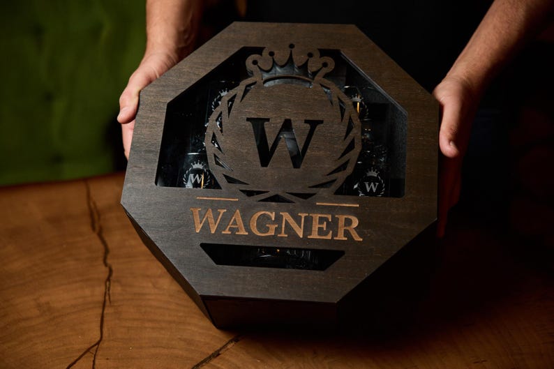 May include: Dark wooden box with an octagonal shape, holding several bottles. The box features a decorative emblem with the letter "W" and the name "Wagner" in a copper color. The box is held by a person.