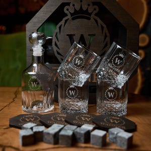 May include: A clear glass decanter with a stopper, three engraved whiskey glasses, coasters, and whiskey stones. The items are branded with a crown and the letter "W" and the name "Wagner". The set is displayed on a wooden surface.