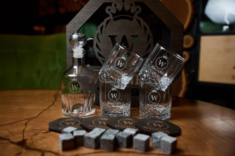 May include: A clear glass decanter with a stopper, three engraved whiskey glasses, coasters, and whiskey stones. The items are branded with a crown and the letter "W" and the name "Wagner". The set is displayed on a wooden surface.
