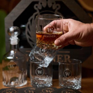 May include: A set of clear whiskey glasses and a decanter with the name "WAGNER" etched on them. One glass is held up, filled with amber liquid. The glasses have a textured base and are part of a gift set.