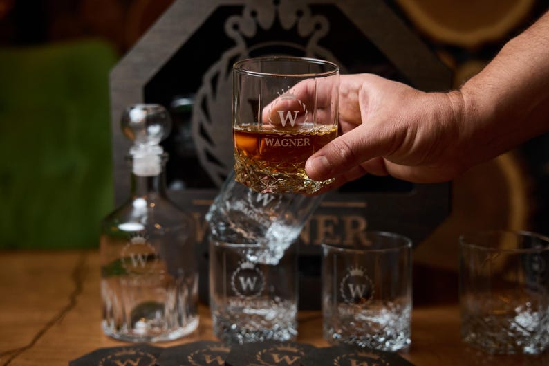May include: A set of clear whiskey glasses and a decanter with the name "WAGNER" etched on them. One glass is held up, filled with amber liquid. The glasses have a textured base and are part of a gift set.