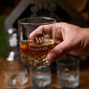 May include: A clear, square-shaped glass with a textured base, filled with amber liquid. The glass is engraved with a crown above the letter "W" and the name "WAGNER". The glass is held by a hand, with other glasses visible in the background.