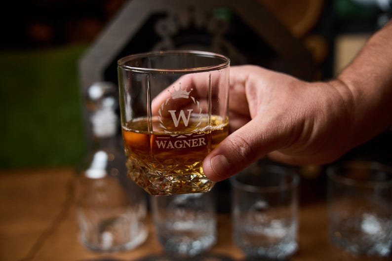 May include: A clear, square-shaped glass with a textured base, filled with amber liquid. The glass is engraved with a crown above the letter "W" and the name "WAGNER". The glass is held by a hand, with other glasses visible in the background.