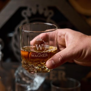 May include: A clear, etched whiskey glass filled with amber liquid, held by a hand. The glass features a crown design with the letter "W" and the name "WAGNER" below. The glass has a textured base.