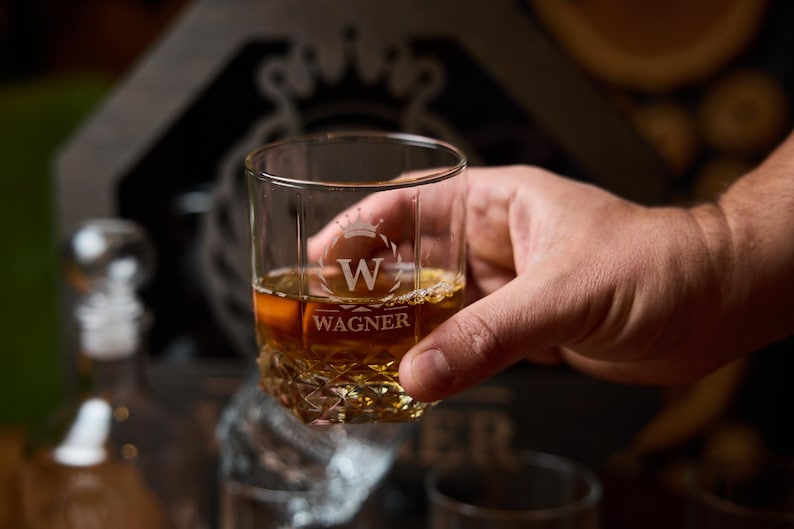 May include: A clear, etched whiskey glass filled with amber liquid, held by a hand. The glass features a crown design with the letter "W" and the name "WAGNER" below. The glass has a textured base.