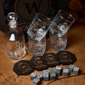 May include: A clear glass decanter with a round stopper, four etched whiskey glasses, coasters, and whiskey stones. The items are arranged on a wooden surface. The coasters and glasses have the word "WAGNER" and a crown logo.