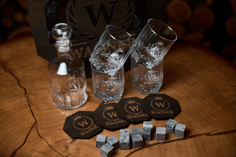May include: A clear glass decanter with a round stopper, four etched whiskey glasses, coasters, and whiskey stones. The items are arranged on a wooden surface. The coasters and glasses have the word "WAGNER" and a crown logo.