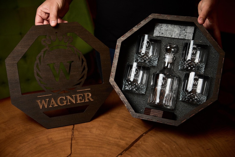May include: A wooden gift set with a decanter, four glasses, and ice stones. The decanter and glasses are clear with a decorative design. The wooden box and lid are dark brown with the name "Wagner" engraved.