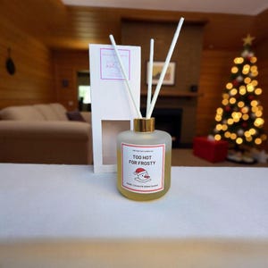 May include: A frosted glass diffuser bottle with a gold cap and white reeds. The label reads "TOO HOT FOR FROSTY" with a snowman graphic. The box behind the bottle has a logo and a window.