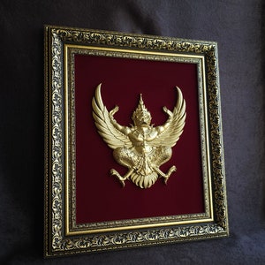 May include: A gold-colored Garuda, the mythical bird-like creature, is framed in a black and gold ornate frame. The frame has a red velvet backing.