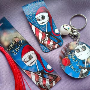 Può includere: Set di tre oggetti con un personaggio dei cartoni animati con un cappello da Babbo Natale. Include due segnalibri con nappe rosse e un portachiavi. I segnalibri e il portachiavi hanno il testo "WHAT'S THIS?" e un design a strisce rosse e bianche.