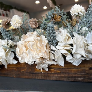 Puede incluir: Un recipiente de madera lleno de un arreglo de flores artificiales. Las flores son principalmente crema, blancas y beige, con acentos de azul verdoso y marr&oacute;n. El recipiente est&aacute; te&ntilde;ido de color marr&oacute;n oscuro.