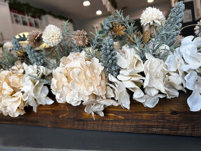 Puede incluir: Un recipiente de madera lleno de un arreglo de flores artificiales. Las flores son principalmente crema, blancas y beige, con acentos de azul verdoso y marr&oacute;n. El recipiente est&aacute; te&ntilde;ido de color marr&oacute;n oscuro.