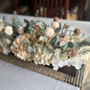 Puede incluir: Centro de mesa rectangular de madera lleno de flores artificiales en tonos crema, beige y verde. El arreglo incluye hortensias, peque&ntilde;as flores redondas y ramitas de vegetaci&oacute;n, sobre un camino de mesa de yute tejido.
