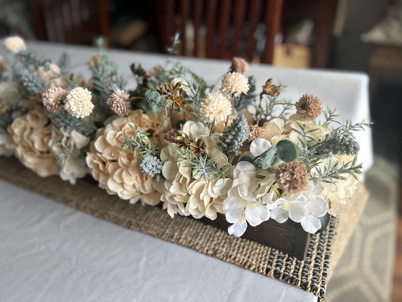 Puede incluir: Centro de mesa rectangular de madera lleno de flores artificiales en tonos crema, beige y verde. El arreglo incluye hortensias, peque&ntilde;as flores redondas y ramitas de vegetaci&oacute;n, sobre un camino de mesa de yute tejido.
