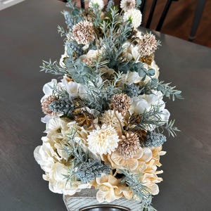Puede incluir: Arreglo floral rectangular de madera con asa de bronce. El arreglo presenta flores crema, beige y marr&oacute;n, acentuadas con follaje verde y verde azulado. Las flores est&aacute;n dispuestas en una caja de madera r&uacute;stica.