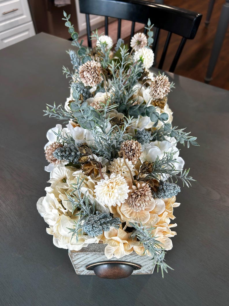 Puede incluir: Arreglo floral rectangular de madera con asa de bronce. El arreglo presenta flores crema, beige y marr&oacute;n, acentuadas con follaje verde y verde azulado. Las flores est&aacute;n dispuestas en una caja de madera r&uacute;stica.