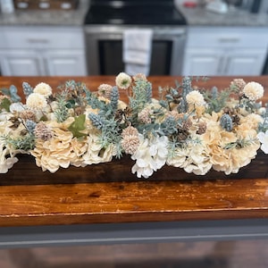 Puede incluir: Una jardinera rectangular de madera llena de flores artificiales. El arreglo incluye hortensias color crema, ramitas azules y verdes y detalles en beige y blanco. La caja est&aacute; sobre una mesa de madera.