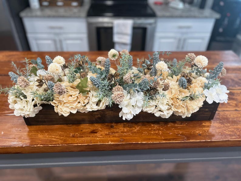 Puede incluir: Una jardinera rectangular de madera llena de flores artificiales. El arreglo incluye hortensias color crema, ramitas azules y verdes y detalles en beige y blanco. La caja est&aacute; sobre una mesa de madera.