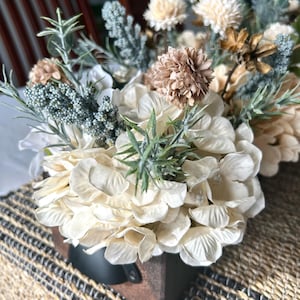 Puede incluir: Arreglo floral en una caja de madera. El arreglo presenta hortensias color crema, flores secas beige y marr&oacute;n, y ramitas de follaje verde y verde azulado. La caja tiene un asa de metal negro.