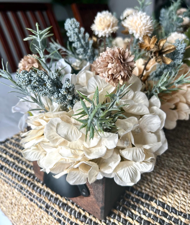 Puede incluir: Arreglo floral en una caja de madera. El arreglo presenta hortensias color crema, flores secas beige y marr&oacute;n, y ramitas de follaje verde y verde azulado. La caja tiene un asa de metal negro.