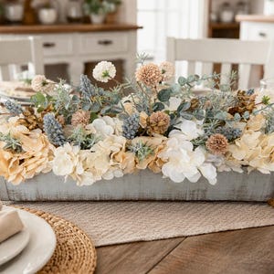 May include: A rectangular wooden centerpiece filled with an assortment of artificial flowers in cream, peach, and blue hues. The arrangement sits on a wooden table, with place settings visible in the foreground.