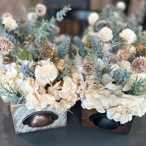 Puede incluir: Dos arreglos florales decorativos en peque&ntilde;as cajas de madera. Las flores son crema, beige y verde apagado, con acentos marrones y azules. Cada caja tiene un tirador de caj&oacute;n de metal. Las cajas est&aacute;n sobre una superficie gris.
