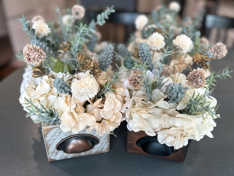 Puede incluir: Dos arreglos florales decorativos en peque&ntilde;as cajas de madera. Las flores son crema, beige y verde apagado, con acentos marrones y azules. Cada caja tiene un tirador de caj&oacute;n de metal. Las cajas est&aacute;n sobre una superficie gris.