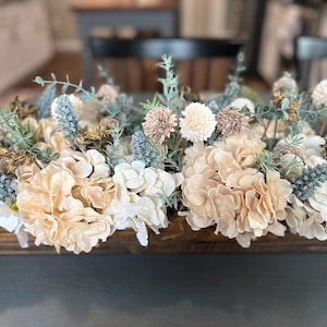Puede incluir: Un centro de mesa de madera lleno de una variedad de flores artificiales en tonos suaves y neutros. El arreglo incluye grandes hortensias cremosas, delicadas flores rosas y blancas y acentos de follaje azul-verdoso. Las flores est&aacute;n dispuestas en una caja de madera rectangular.