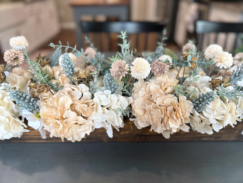 Puede incluir: Un centro de mesa de madera lleno de una variedad de flores artificiales en tonos suaves y neutros. El arreglo incluye grandes hortensias cremosas, delicadas flores rosas y blancas y acentos de follaje azul-verdoso. Las flores est&aacute;n dispuestas en una caja de madera rectangular.
