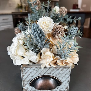 Puede incluir: Arreglo floral en una caja de madera envejecida. El arreglo presenta flores crema, beige y marr&oacute;n con acentos azul-verdes. La caja tiene un asa de metal y un acabado azul claro desgastado, creando una est&eacute;tica r&uacute;stica.