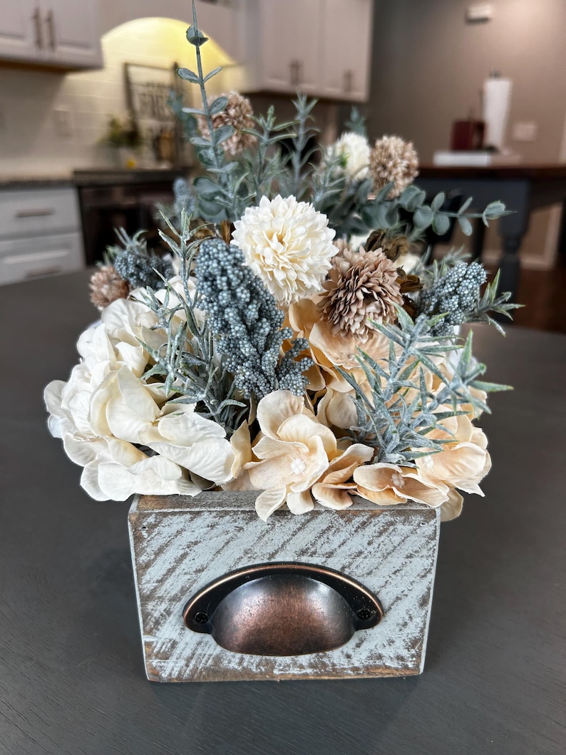 Puede incluir: Arreglo floral en una caja de madera envejecida. El arreglo presenta flores crema, beige y marr&oacute;n con acentos azul-verdes. La caja tiene un asa de metal y un acabado azul claro desgastado, creando una est&eacute;tica r&uacute;stica.