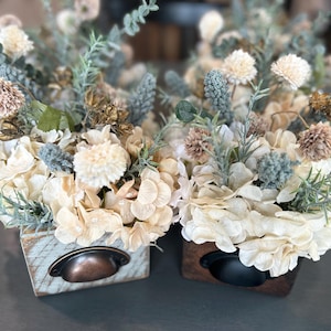Puede incluir: Dos arreglos florales decorativos en peque&ntilde;as cajas de madera. Las flores son crema, beige y verde apagado, con acentos azul-gris. Cada caja tiene un tirador de caj&oacute;n de metal. La est&eacute;tica general es r&uacute;stica y elegante. Las cajas miden unos 15 cm de ancho.