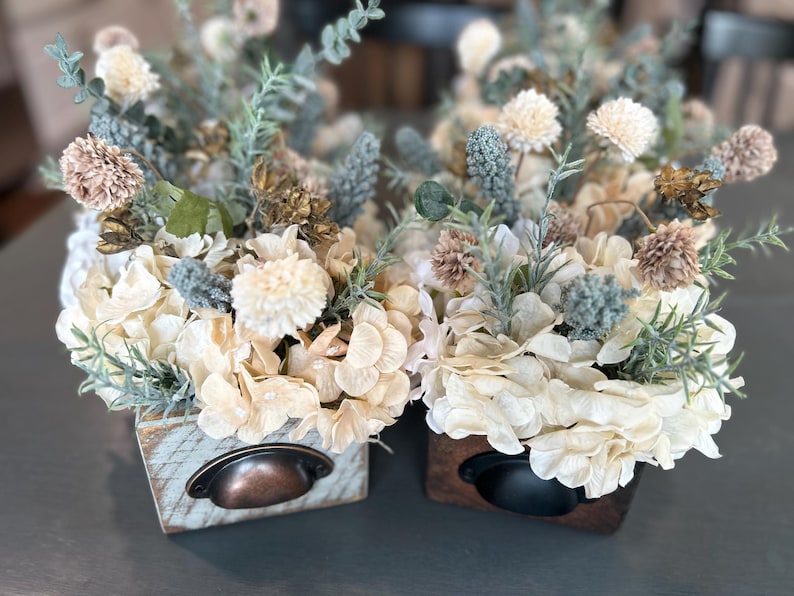 Puede incluir: Dos arreglos florales decorativos en peque&ntilde;as cajas de madera. Las flores son crema, beige y verde apagado, con acentos azul-gris. Cada caja tiene un tirador de caj&oacute;n de metal. La est&eacute;tica general es r&uacute;stica y elegante. Las cajas miden unos 15 cm de ancho.