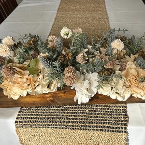 Puede incluir: Centro de mesa de madera lleno de flores y vegetaci&oacute;n artificiales crema, blancas y beige. El arreglo incluye hortensias y otros elementos florales. El centro de mesa est&aacute; sobre un mantel blanco con un camino de mesa tejido.