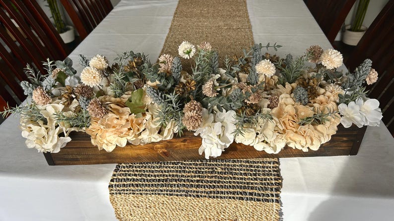 Puede incluir: Centro de mesa de madera lleno de flores y vegetaci&oacute;n artificiales crema, blancas y beige. El arreglo incluye hortensias y otros elementos florales. El centro de mesa est&aacute; sobre un mantel blanco con un camino de mesa tejido.
