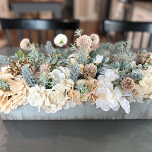 Puede incluir: Un jardinera rectangular de madera llena de flores artificiales. El arreglo incluye hortensias crema, blancas y azul claro, junto con otras plantas verdes y flores esf&eacute;ricas de color canela. La jardinera tiene un acabado azul claro envejecido, de unos 30 cm de largo.