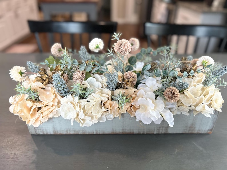Puede incluir: Un jardinera rectangular de madera llena de flores artificiales. El arreglo incluye hortensias crema, blancas y azul claro, junto con otras plantas verdes y flores esf&eacute;ricas de color canela. La jardinera tiene un acabado azul claro envejecido, de unos 30 cm de largo.