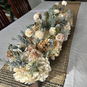 Puede incluir: Un arreglo floral central con flores artificiales crema, melocot&oacute;n y verde salvia. El arreglo se coloca sobre un camino de mesa tejido, creando una est&eacute;tica natural y elegante. Las flores incluyen hortensias y otras flores decorativas.