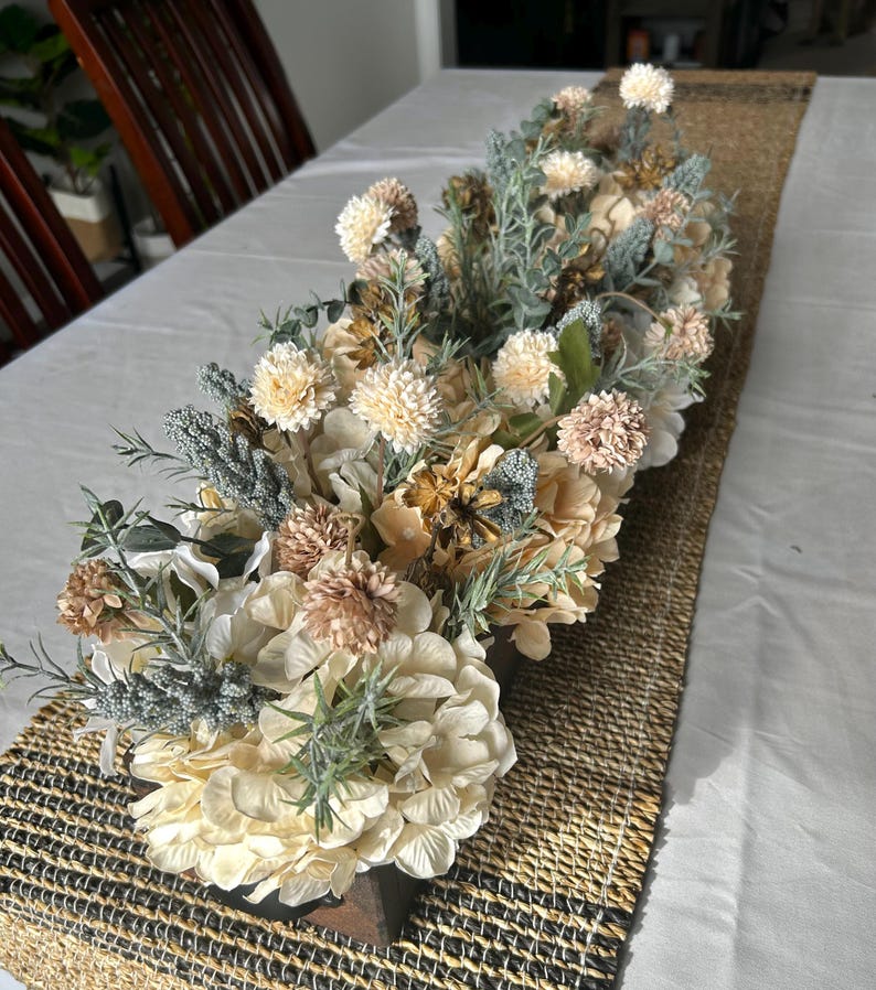 Puede incluir: Un arreglo floral central con flores artificiales crema, melocot&oacute;n y verde salvia. El arreglo se coloca sobre un camino de mesa tejido, creando una est&eacute;tica natural y elegante. Las flores incluyen hortensias y otras flores decorativas.