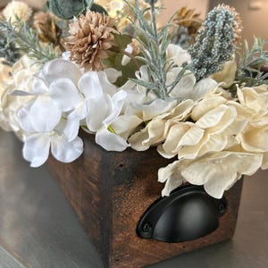 Puede incluir: Una caja rectangular de madera llena de flores artificiales. Las flores son principalmente blancas y crema, con acentos verdes y marrones. La caja tiene un acabado marr&oacute;n oscuro y un asa de metal negro.