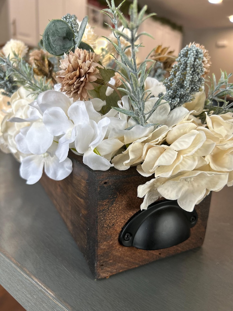 Puede incluir: Una caja rectangular de madera llena de flores artificiales. Las flores son principalmente blancas y crema, con acentos verdes y marrones. La caja tiene un acabado marr&oacute;n oscuro y un asa de metal negro.
